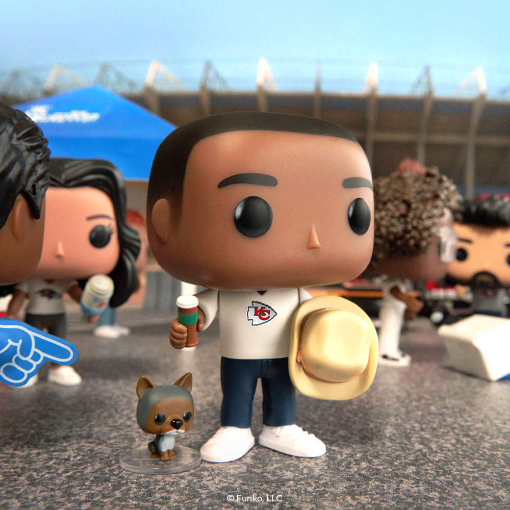 This lifestyle photo shows a POP! Yourself collectible figure of a Chiefs fan. He's holding a cowboy hat. There is a stadium in the background, along with other POP! Yourself figures and toy cars to give the impression of a tailgating party.